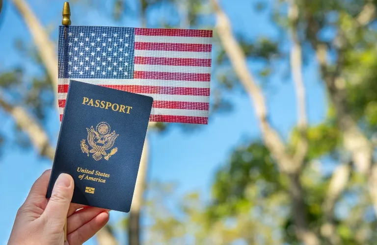 Hand holding blue United States passport and small American flag outdoors with blurred trees background.
