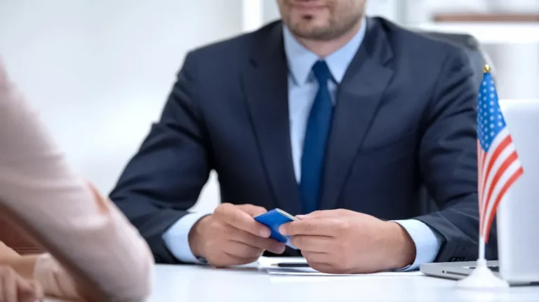 Man in navy blue suit holding a blue passport, small American flag on desk, laptop partially visible.